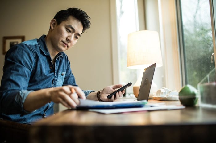 A person flips through papers at a home desk with a phone and laptop.