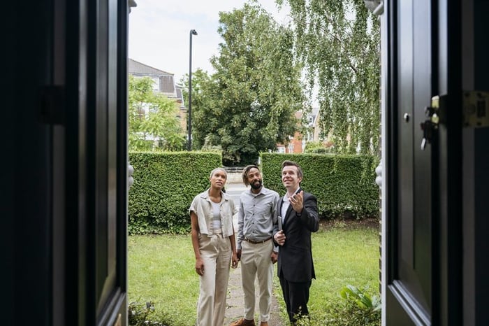 A smiling real estate agent shows two clients features of a home's exterior.