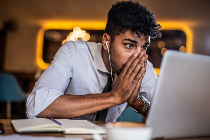 A person wearing headphones puts their hands over their mouth while looking at a laptop screen in a surprised manner.