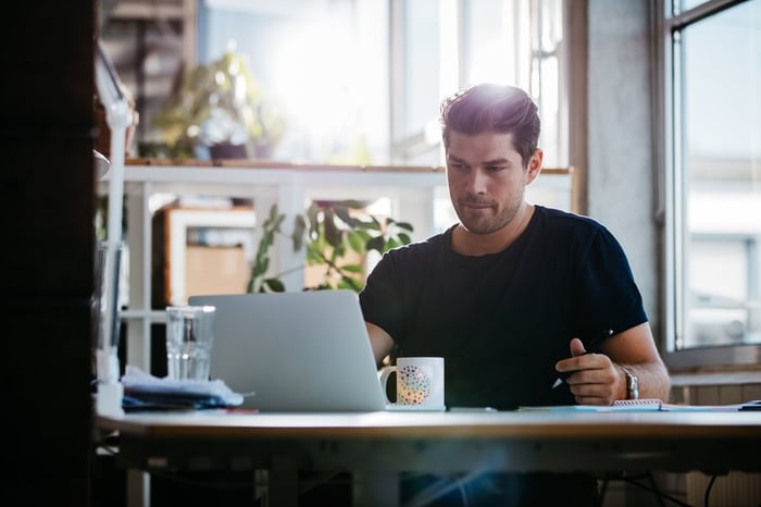 A man sitting at a sunny desk and looking up something on a laptop.