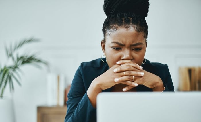 A young business professional worriedly looks at a computer screen as their head rests on their clasped hands.