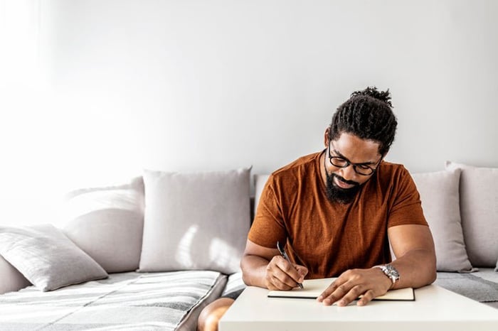 A man sitting on his couch in his sunny living room and writing in a notebook.