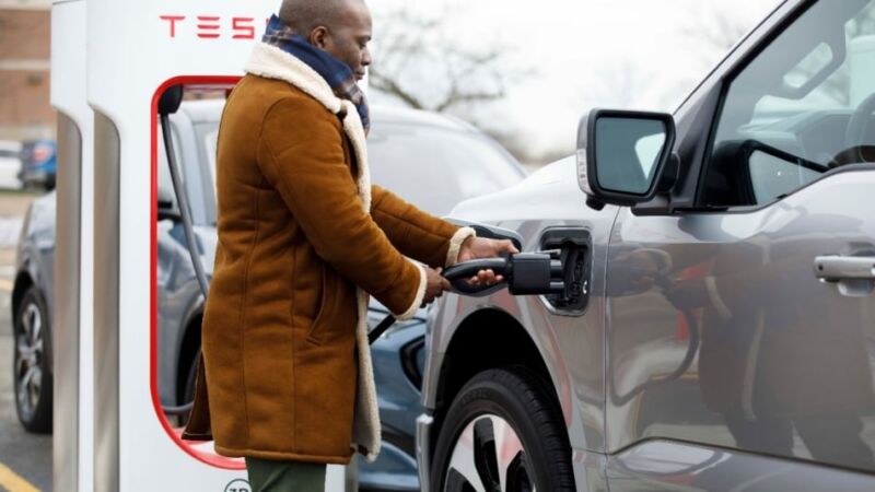 A man plugs a silver Mustang Mach-E into a Tesla Supercharger