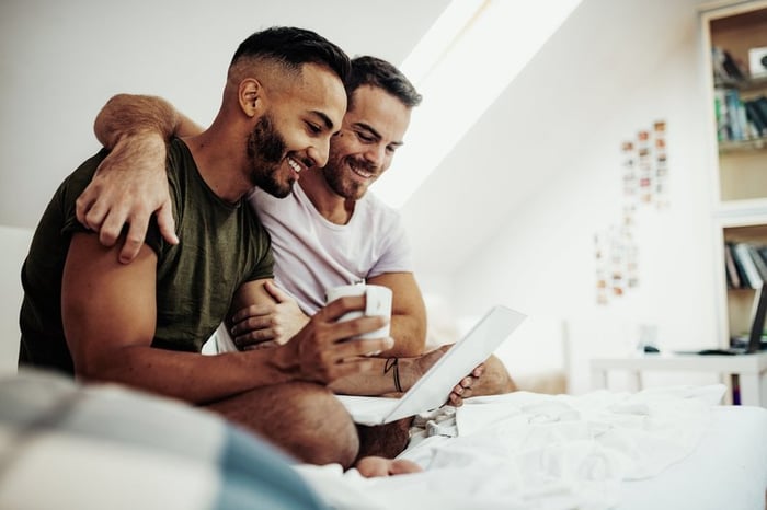 A couple sits in bed drinking coffee and smiling at something on a laptop screen.