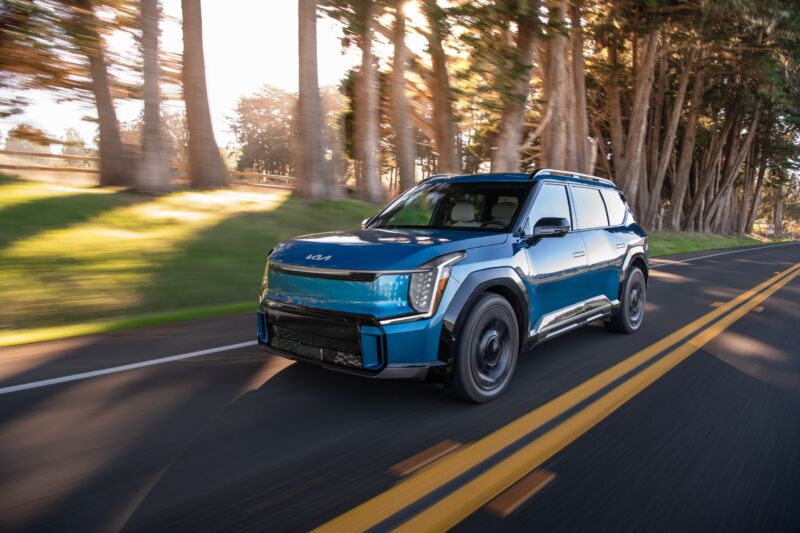 A blue Kia EV9 drives along a forest road in California