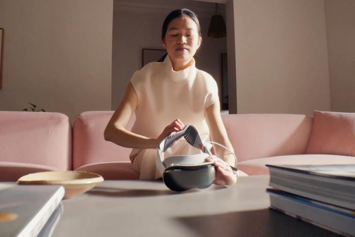 A person picks up an Apple Vision Pro headset that is resting on a desk.