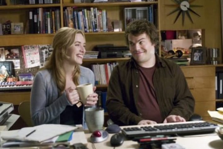 A woman holding a coffee cup sits down to a man at a keyboard.