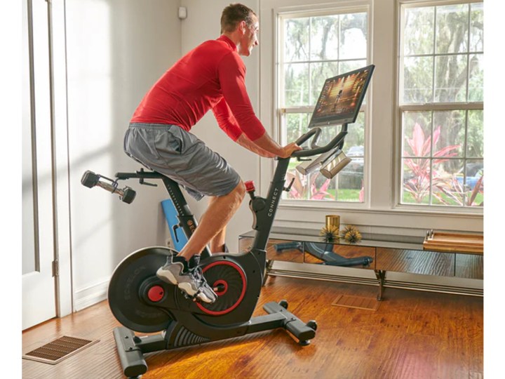 Riding an Echelon Connect Bike EX-5s-22 inside a house with a view of flowering bushes.