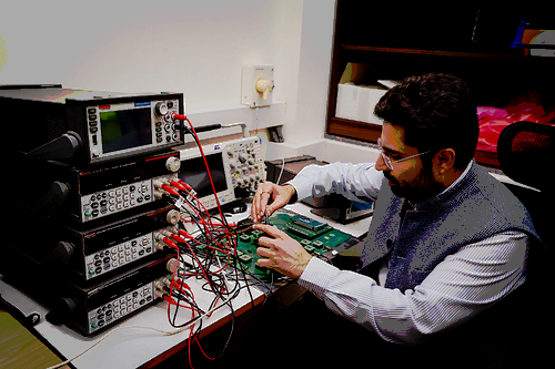 A man is sitting at a table working on a circuit board and four machines that have red and black wires inserted into them. 