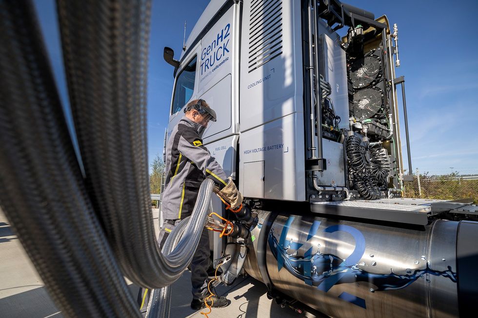 A person in protective hood gear and thick gloves connects long, thick pipes to the front of a semi truck, where a tank says H2.