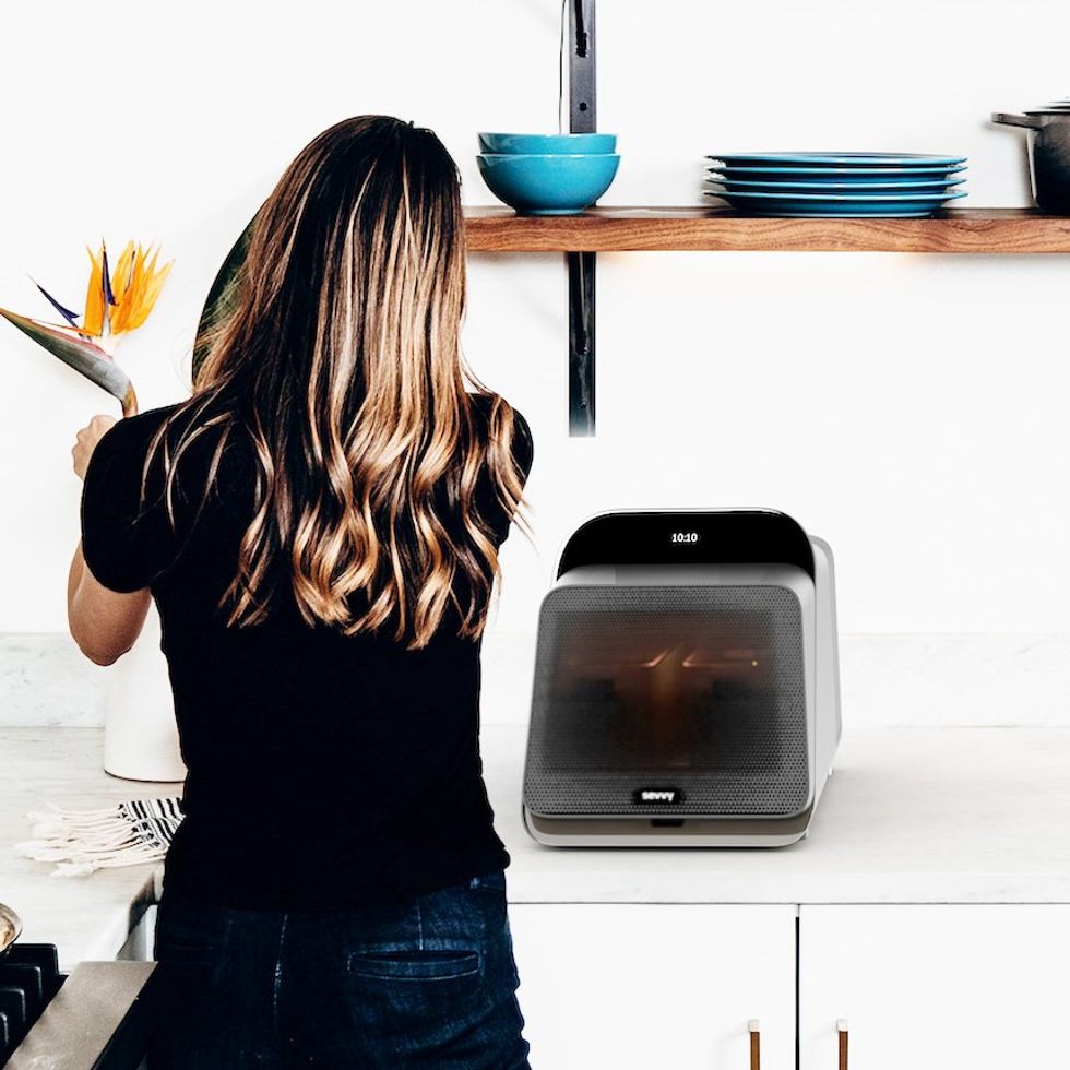 A woman in a kitchen facing away from camera, a square appliance slightly bigger than a breadbox on the counter.