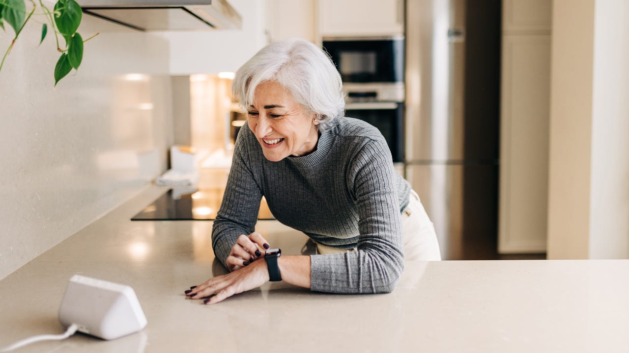 Senior woman smiling happily while using smart devices in her kitchen. Cheerful elderly woman using a home assistant to perform tasks at home.