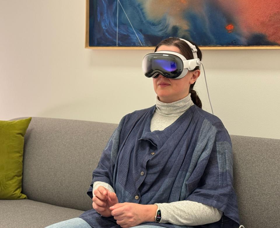 A woman with dark hair wearing the Apple Vision Pro headset, sat back on a gray couch.