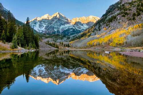 Sunrise over the iconic Maroon Bells mountains near Aspen, Colorado. Clear sky above with colorful aspen trees in full fall foliage color splendor with the entire scene reflected in the small lake in the foreground.