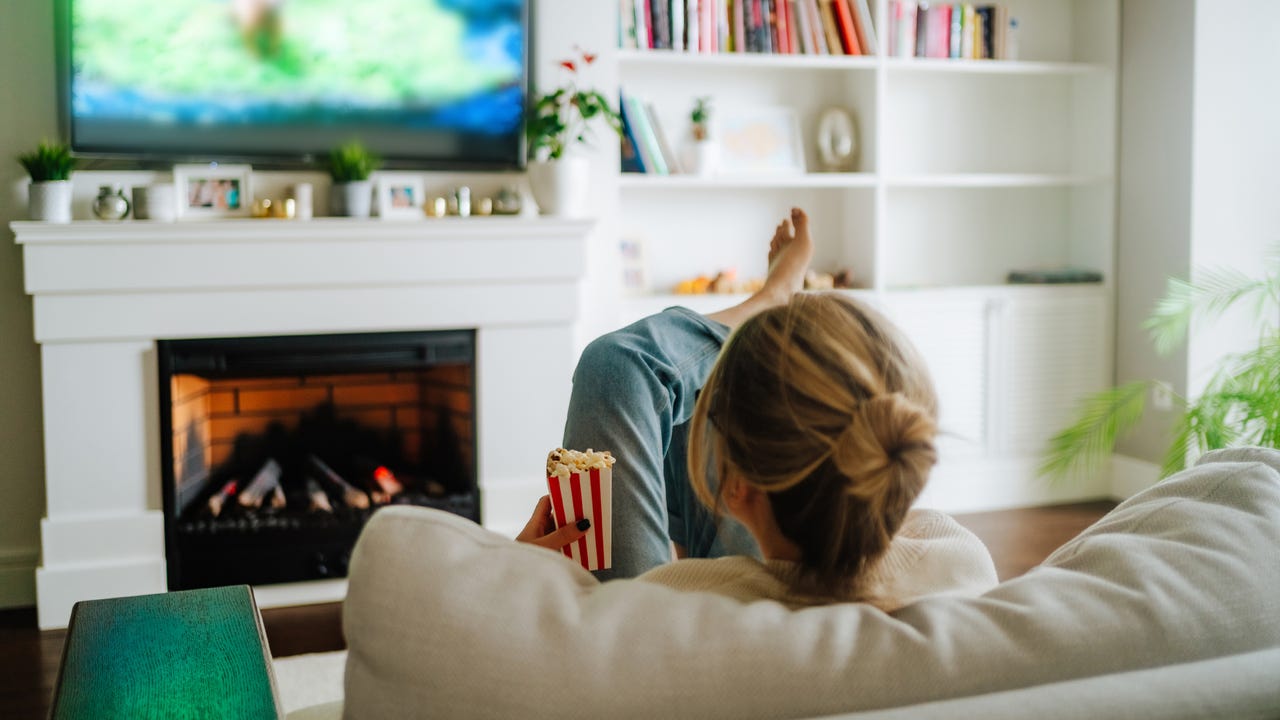 Blond woman with hair bun watching TV on a cozy sofa against fireplace indoors