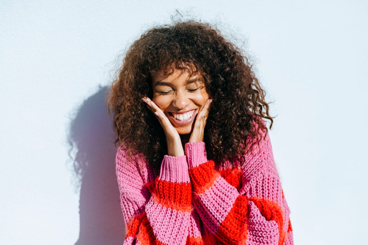 Young woman with curling hair laughing while standing against a wall.