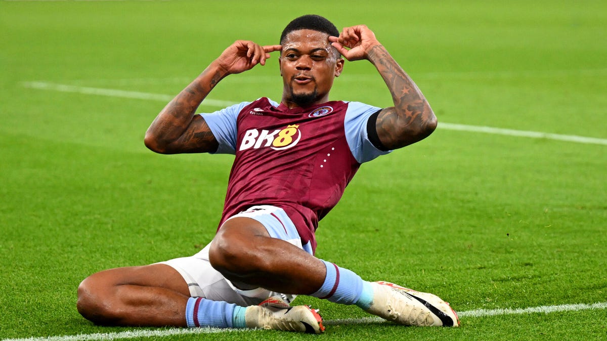 Aston Villa forward Leon Bailey celebrating, sliding on his knees, pointing to his head.