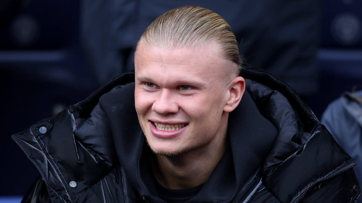 Manchester City striker Erling Haaland smiling, looking to his right, wearing a black coat.