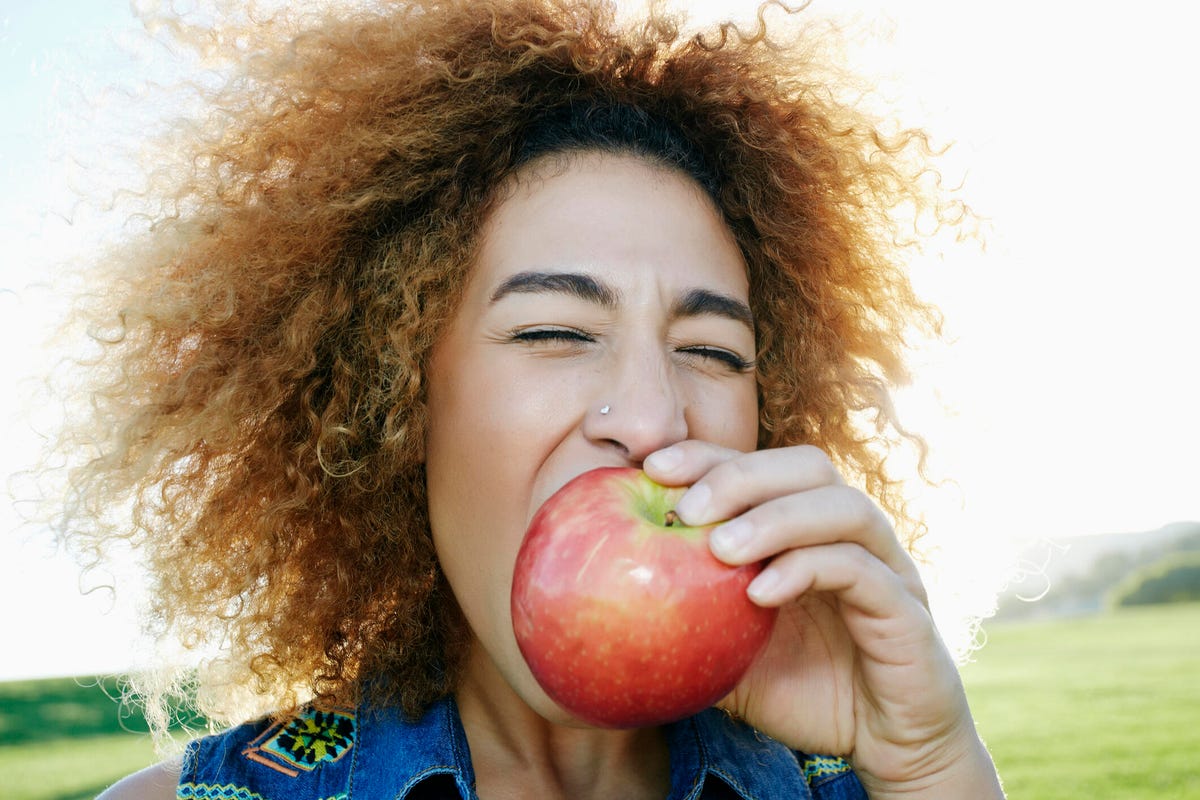 woman with brown curly hair eating a pink apple outside