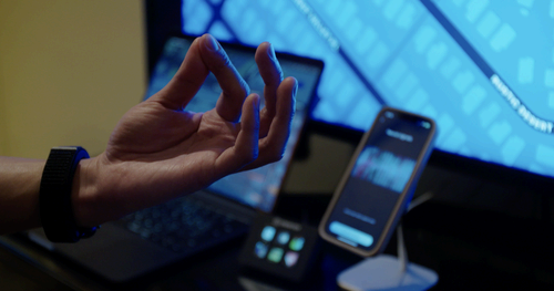 Close up of wrist shows a user wearing the Mudra while pinching their fingers. In the background, connected devices show the neural activity triggered by the hand gesture.