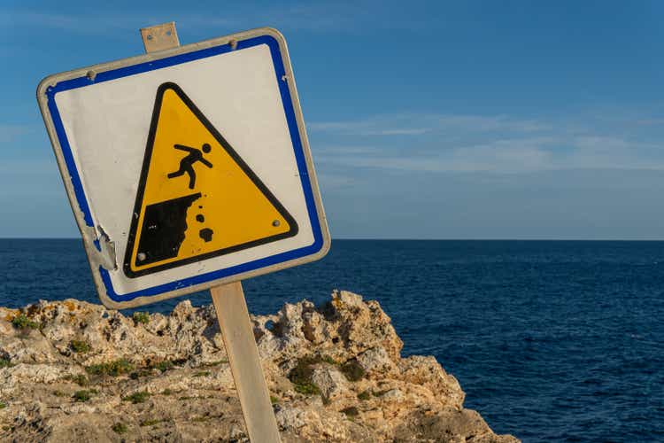 Close-up of a danger sign for people falling down a ravine, crooked. In the background the Mediterranean Sea on the island of Mallorca at sunset