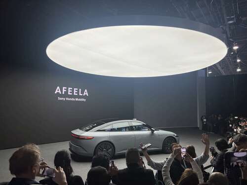 A silver eletric vehicle is displayed on stage under a huge overlight. The car is surrounded by media taking picutres of it.