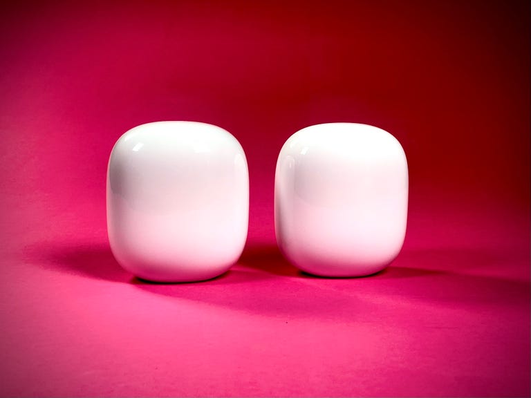 Two white Nest Wifi Pro mesh routers siting side-by-side against a pink background.