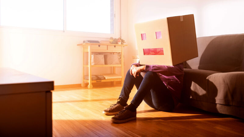 A lazy robot (a man with a box on his head) sits on the floor beside a couch.