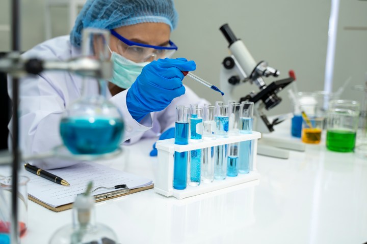 Scientist in laboratory working with an eyedropper, test tubes, and blue liquid and surrounded by various lab equipment.