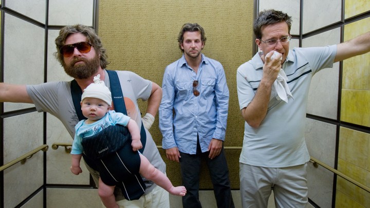 Three men in an elevator, one leaning back one with a cloth to his mouth, the other holding a baby in a carrier in a scene from The Hangover.