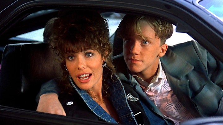 A young teen driving a car, his arm around a gorgeous woman in a scene from Weird Science.