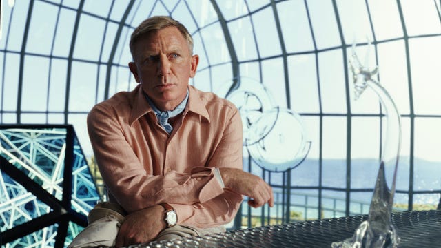 Benoit Blanc, played by Daniel Craig, looks askance with his arms cross inside a large glass atrium