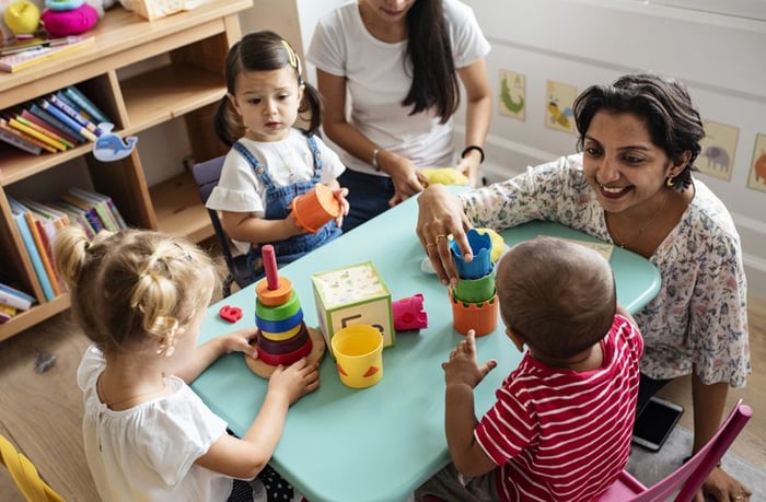 A smiling teacher sitting at a table in her daycare classroom and playing with three young children.