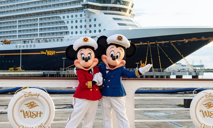 Mickey and Minnie Mouse waving to the camera in front of a Disney cruise ship.