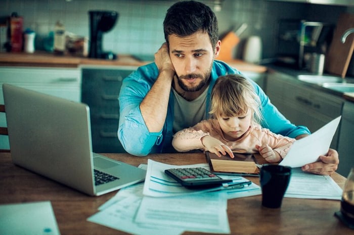 A parent sitting at the kitchen table with paperwork, a calculator, a laptop, and their toddler on their lap.