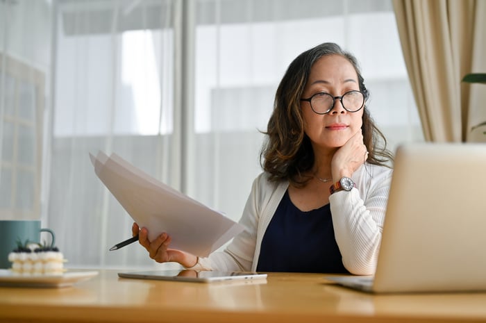 A businessperson looking at an open laptop and holding paperwork in their right hand while seated at a desk.