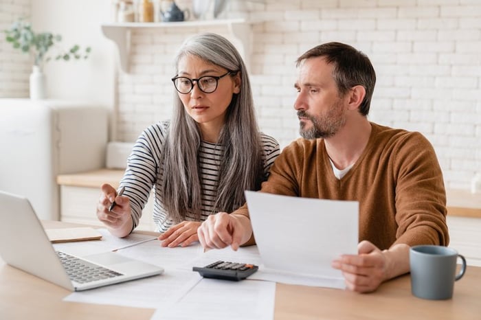 Two people sitting at their kitchen table with an open laptop, paperwork, and a calculator.