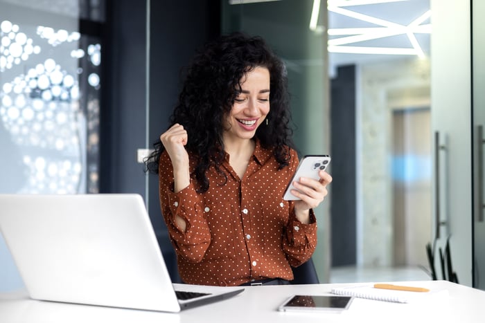 A person sitting at a desk behind a computer while staring at their phone and smiling.