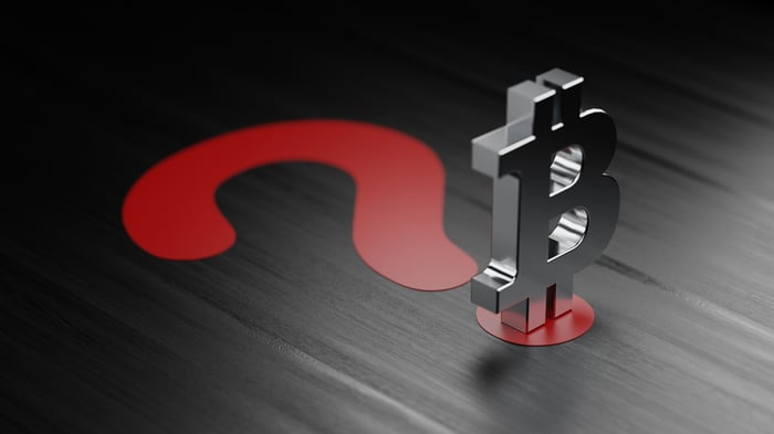 Silver 3D Bitcoin logo with its shadow forming a red question mark on a dark wooden table.