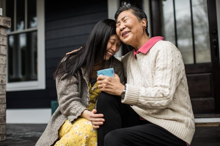 An adult woman and her senior mom sitting on their front porch steps while smiling and hugging.