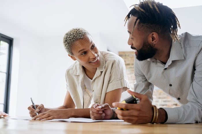 A couple fills out financial paperwork as they smile while leaning against a kitchen island.