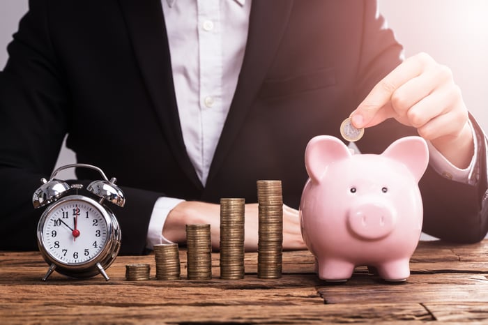 A person puts a coin in a piggy bank alongside a stock of coins and a clock.