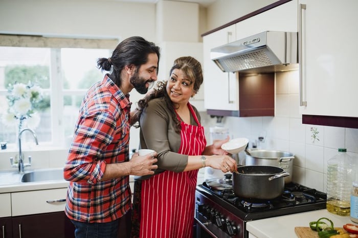 A mother cooking at the stove with her adult son looking over her shoulder.