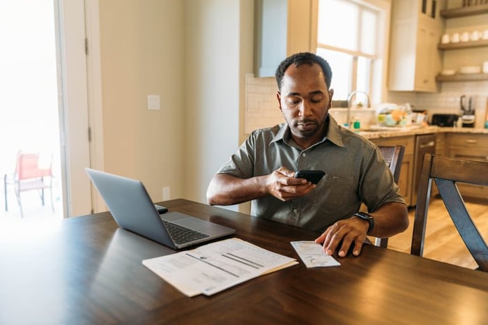 Man sits at counter with phone on laptop utilizing online banking.