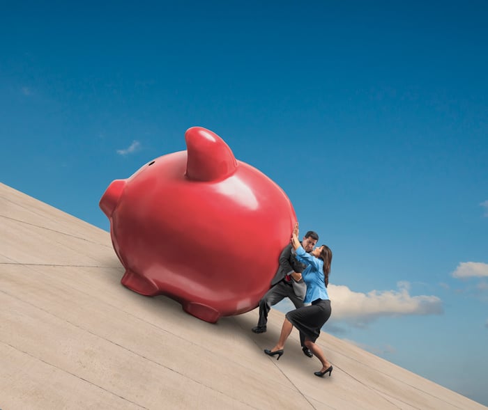 Two people pushing a huge piggy bank up an incline.