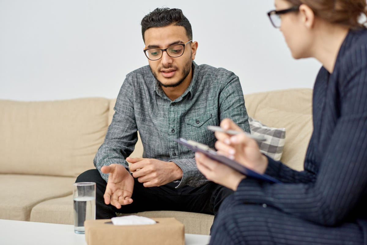 Man sitting on a couch in therapy, talking to his therapist who has a clipboard in her hand.