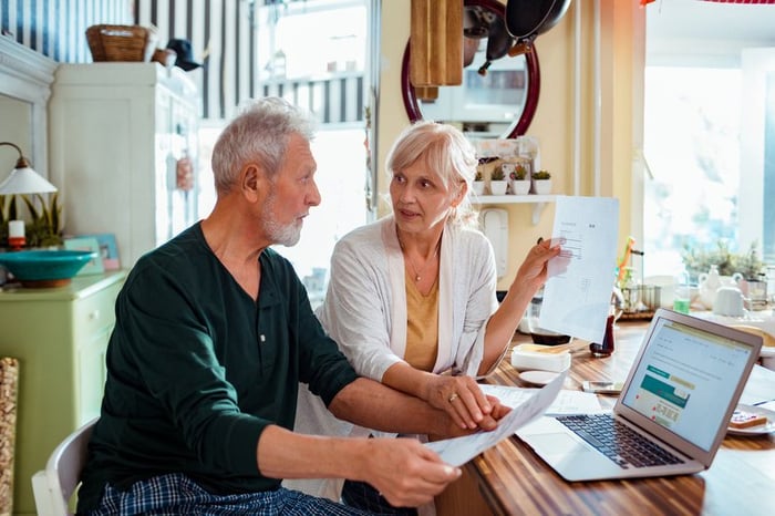 An older couple pays bills at the kitchen table using a laptop.