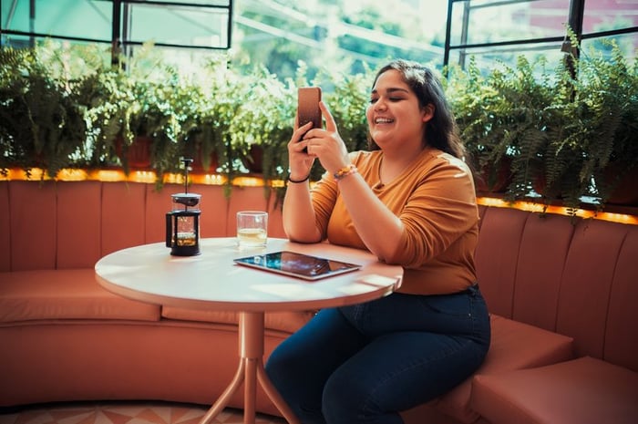 A smiling person sitting at a table in a coffee shop and looking at a phone in their hand