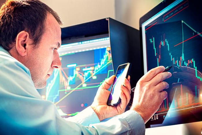 A money manager using a smartphone and stylus to analyze a stock chart displayed on a computer screen.