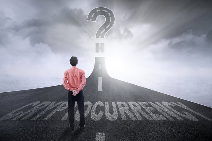 A person stands on a highway marked Cryptocurrency, staring at a giant question mark in the sky.